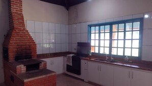 Una cucina con mobili bianchi, un forno a mattoni, una finestra con infissi blu e un pavimento piastrellato.