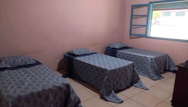 Une chambre avec trois lits, chacun recouvert d’une couvre-lit à motifs, une fenêtre avec un cadre bleu et un sol carrelé.