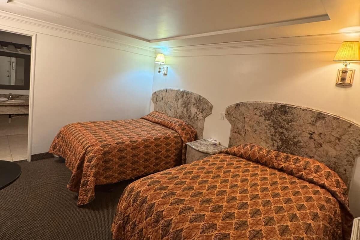 Quarto de hotel com duas camas, cabeceira de mármore, mesa de cabeceira pequena e uma luminária fixada na parede.
