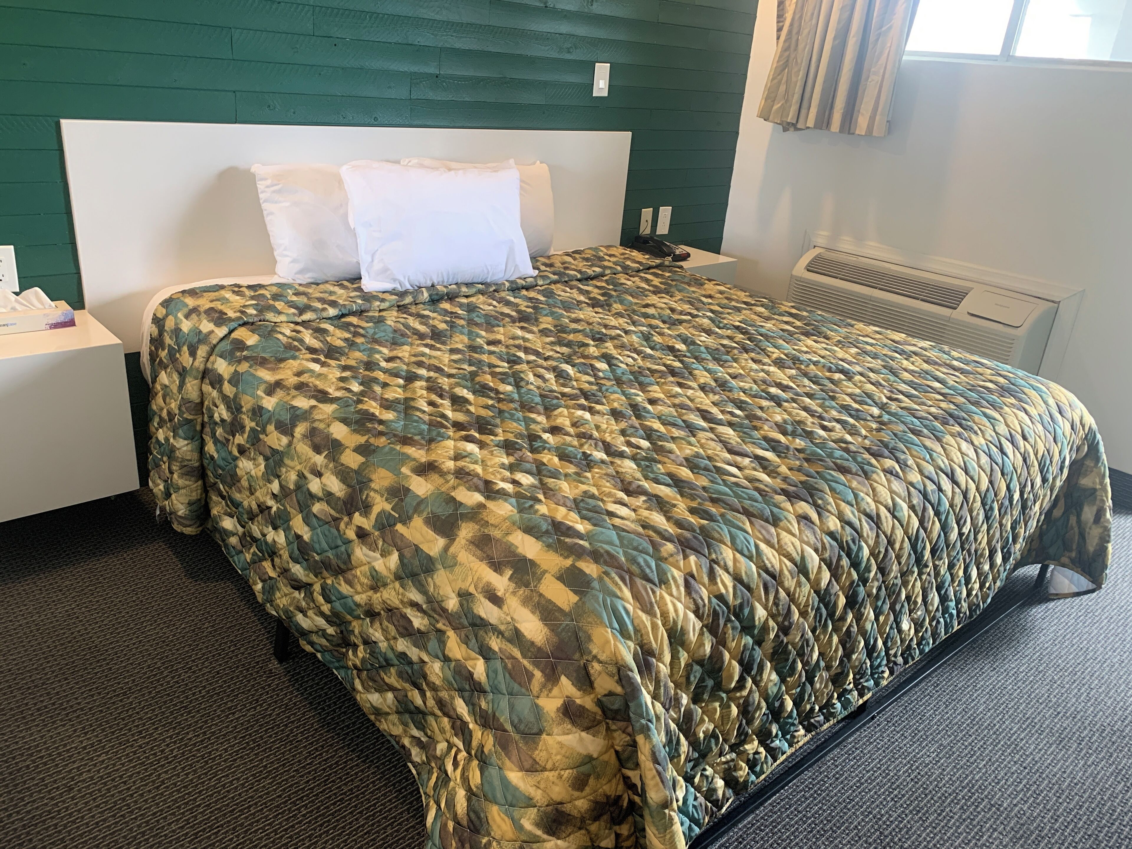 A neatly made bed with a patterned quilt, a white headboard, a bedside table with a phone, and a window with curtains.