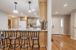 A modern kitchen with wooden cabinets, a marble countertop, and bar stools. - Spacious Retreat: Hot Tub, Game Room & Theater! (Idaho Falls)