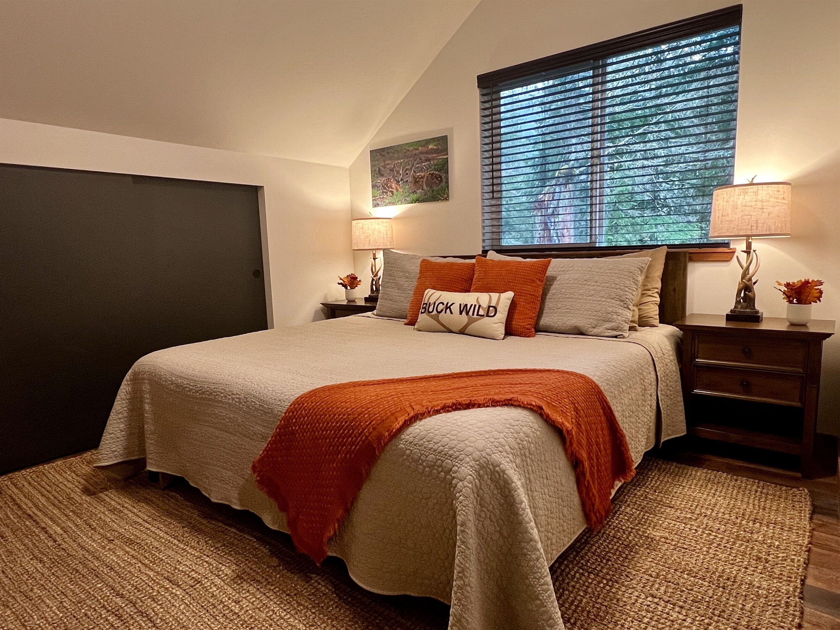 A bedroom with a large bed, a window with blinds, a nightstand with a lamp, and a wall-mounted painting.