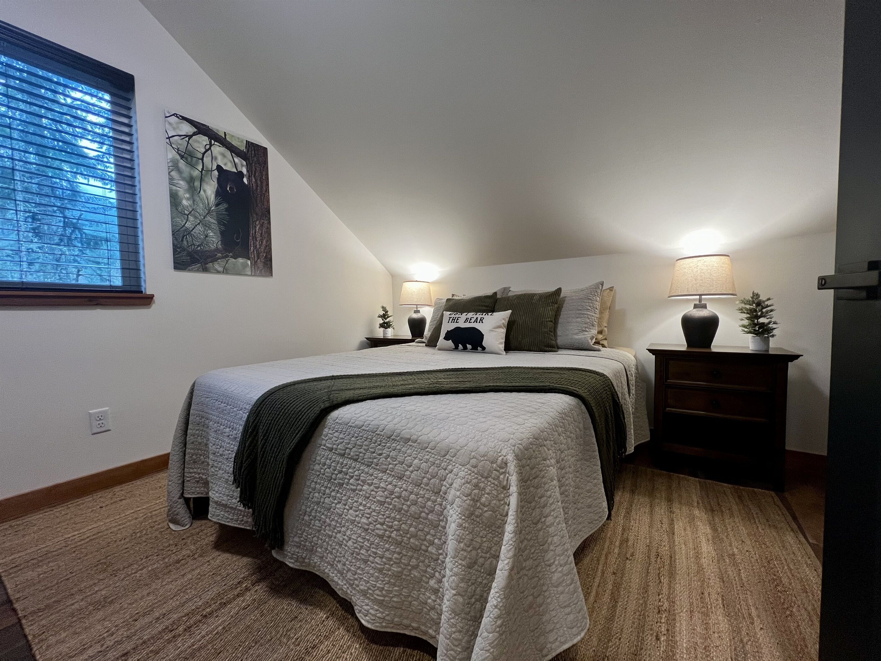 A bedroom with a bed, two bedside tables, a nightstand, a window with blinds, a wall-mounted lamp, and a small potted plant.