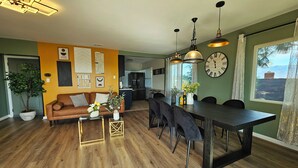 A modern living room with a sofa, coffee table, and a dining area with a table and chairs. There is a large clock on the wall and a view of the outdoors through a window. - Ideally located, easy access, & stunning city views! with king size bed. (Alhambra)