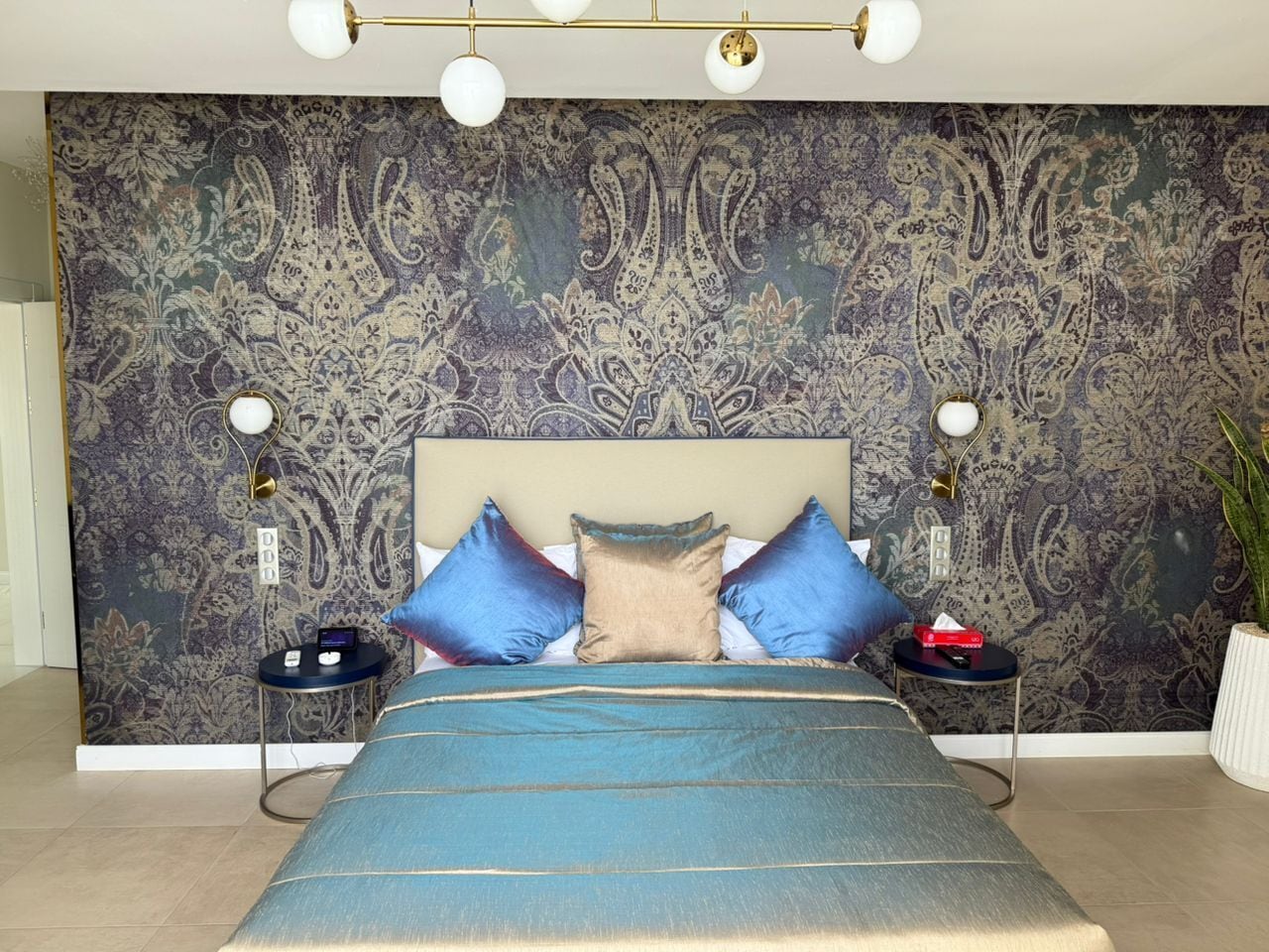 A bedroom with a large bed, a patterned wallpaper, two bedside tables with lamps, and a potted plant.
