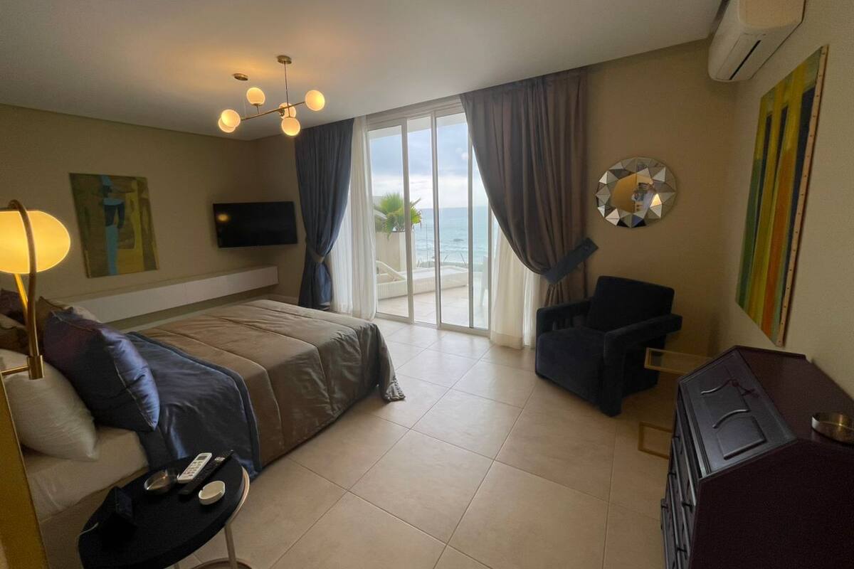 Una habitació d'hotel amb un llit, una taula, una cadira, un televisor i vista a l'oceà a través de grans finestres.