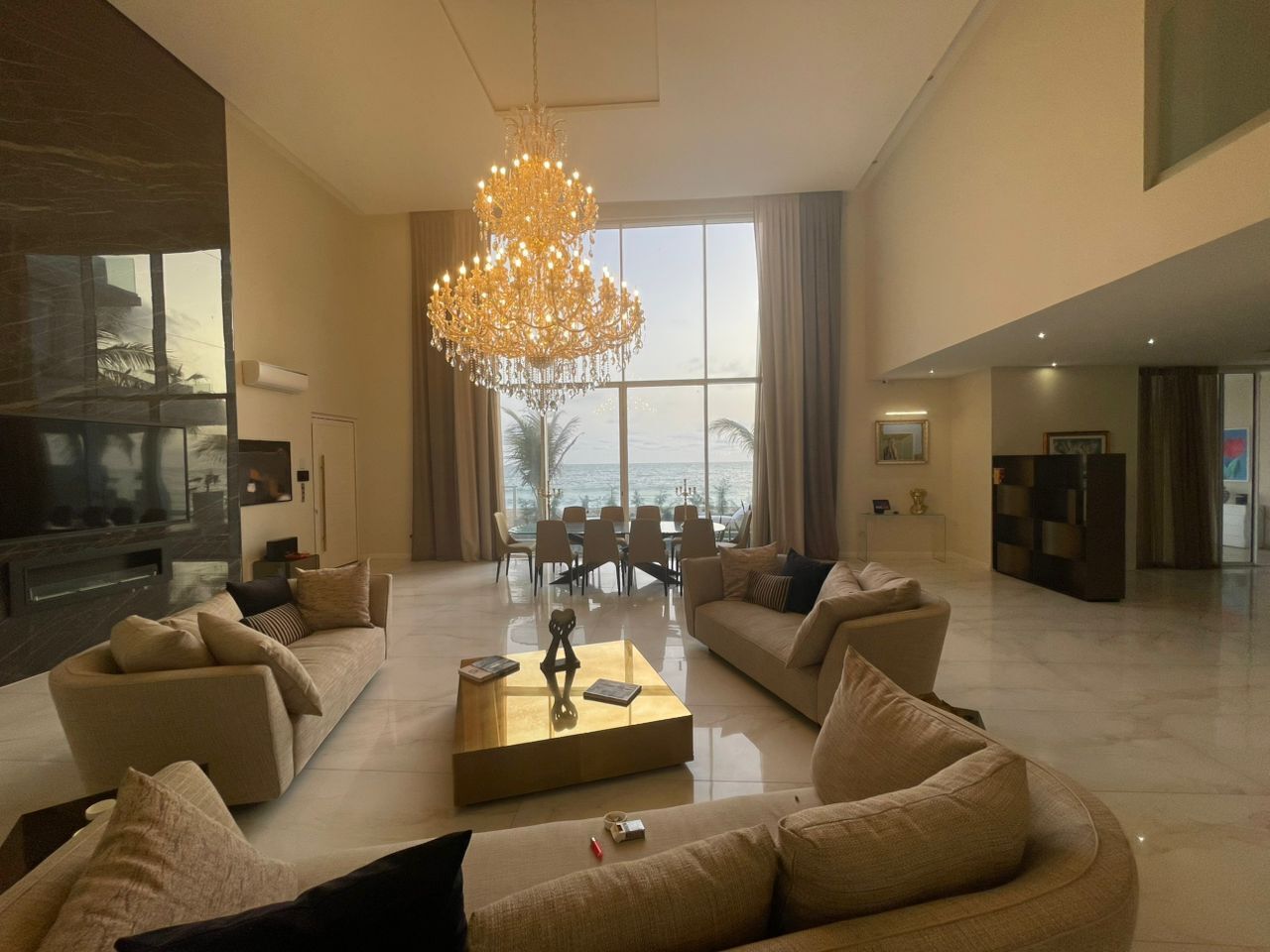 A spacious living room with a large chandelier, a flat-screen TV, and a dining area with a view of the ocean.