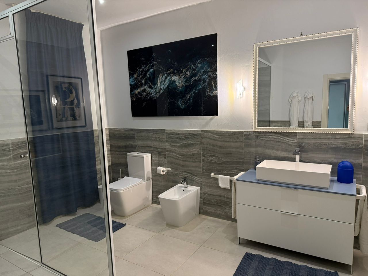 A modern bathroom with a glass shower enclosure, a bidet, a toilet, a sink with a mirror, and a large abstract painting on the wall.
