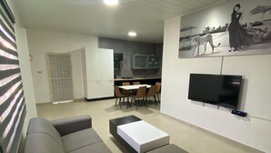 Ein Wohnzimmer mit einer Couch, einem Couchtisch und einem an der Wand montierten Fernseher. Im Hintergrund ist ein Essbereich mit Stühlen und einem Tisch zu sehen.