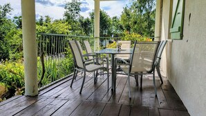 A covered patio with a table and chairs, surrounded by a metal railing, overlooking a garden with various plants and trees.
