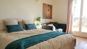 A bedroom with a large bed, a bedside table with a lamp, a chair, a potted plant, and a painting on the wall.