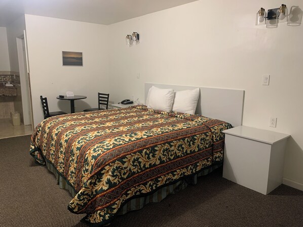 Une chambre à coucher bien rangée, avec une couvre-lit à motifs, une table de chevet, une chaise, une petite table avec un téléphone et un luminaire fixé au mur.