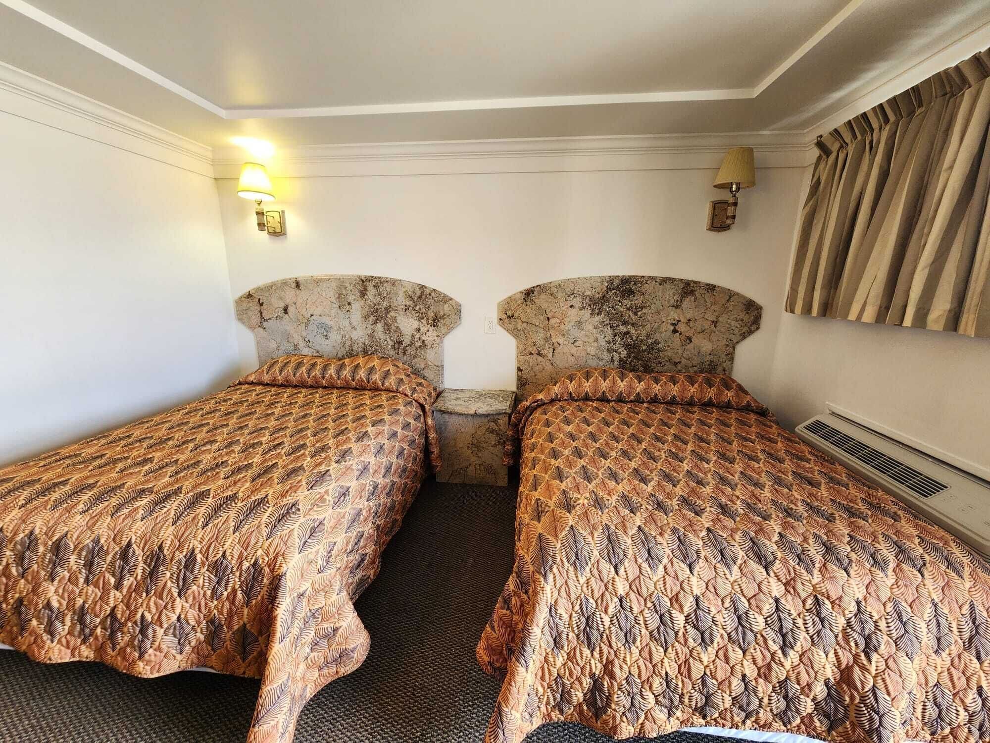 A hotel room with two single beds, each with a patterned bedspread. There are wall-mounted lamps and a heater on the wall.