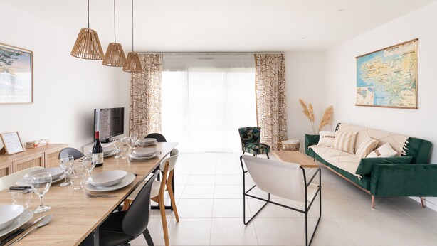 Ein modernes Wohnzimmer mit Essbereich, ausgestattet mit einem grünen Sofa, einem Holztisch und hängenden Rattanlampen.