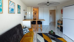 A modern living room with a sofa, a dining area with stools, and a kitchen in the background.