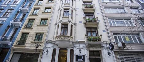A multi-story building with a classic architectural style, featuring balconies with plants, large windows, and a prominent entrance with a sign.
