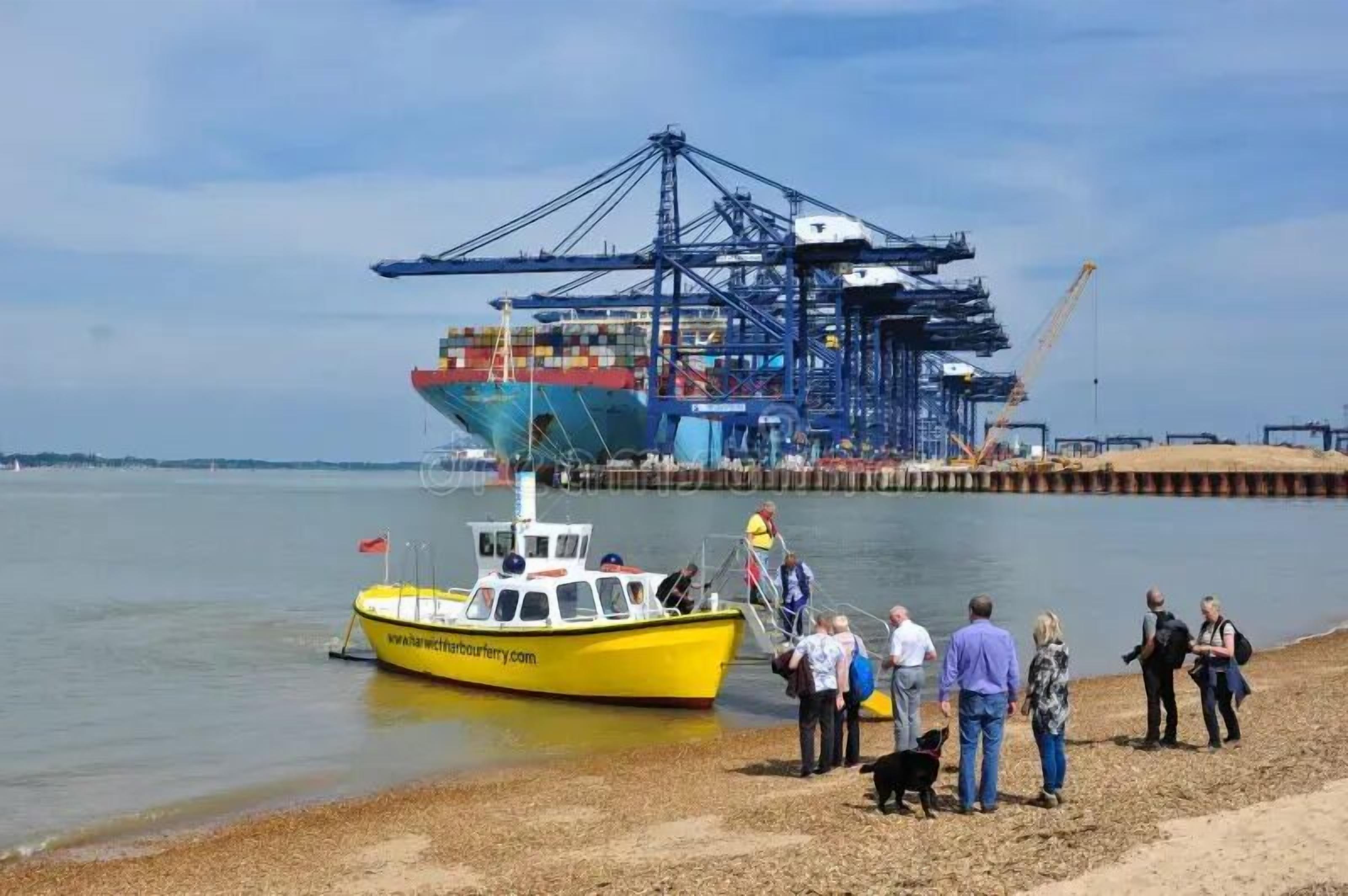 A ferry dock with a yellow boat, a large cargo ship, and a pier with cranes.