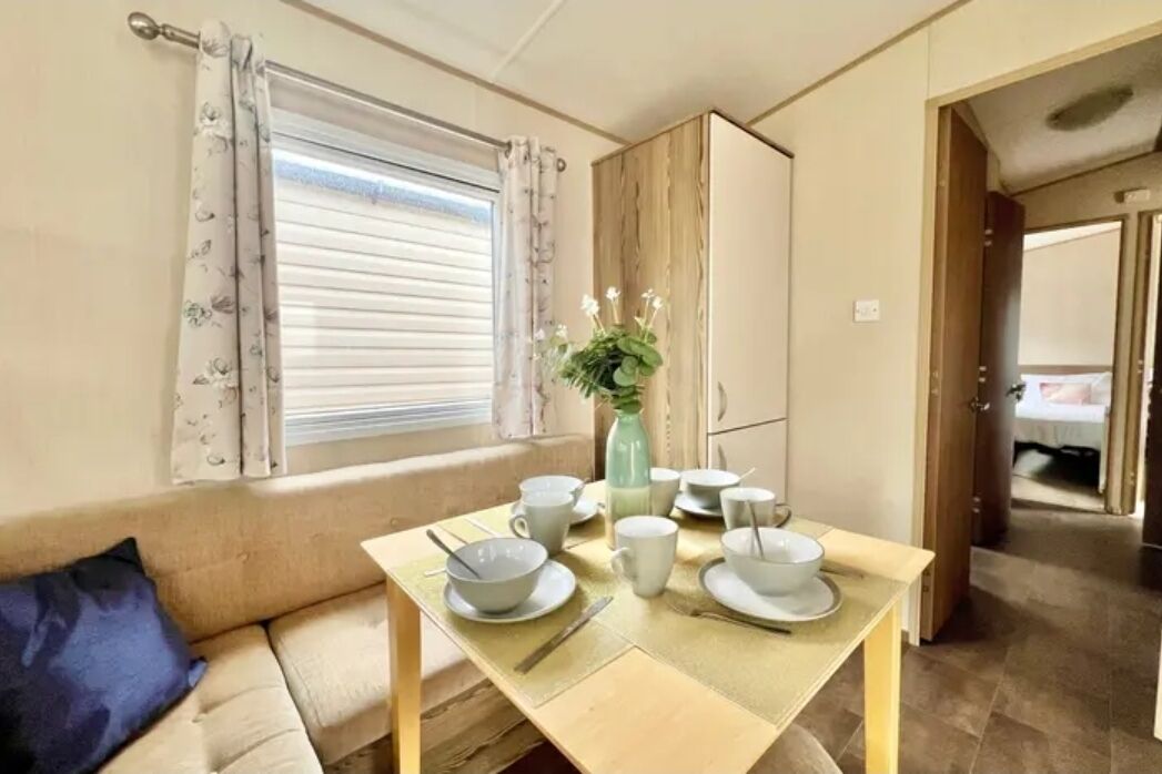 A room with a dining table set for four, a sofa with a blue pillow, a window with curtains, and a wooden cabinet.