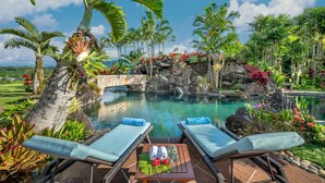Un espace aménagé au bord de la piscine, avec des chaises longues, une table avec des serviettes et un panier de fruits, le tout entouré de plantes tropicales et agrémenté d'une cascade rocheuse en arrière-plan.