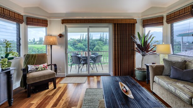 A room with a wooden floor, a sofa, a chair, a table, a lamp, and a view through a sliding glass door to an outdoor area with a table and chairs. - Hanalei Bay Villa 8 (Princeville)
