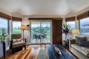 Ein Zimmer mit Holzboden, einer Couch, einem Sessel, einem Tisch, einer Lampe und Blick durch eine Schiebetür auf einen Außenbereich mit Tisch und Stühlen.