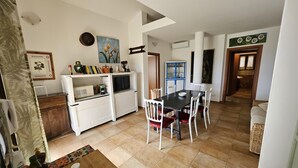 Et moderne køkken med hvide skabe, et spiseområde med et sort bord og hvide stole, og en opholdsstue med en sofa og et mønstret tæppe.