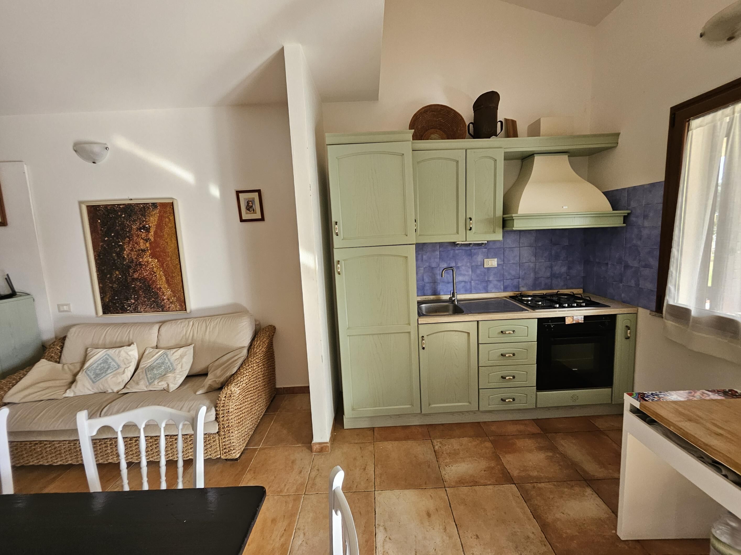 Une cuisine compacte avec des meubles verts, une cuisinière et un évier. On y trouve un coin repas avec une table et des chaises, ainsi qu’un canapé avec des coussins.