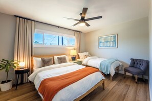 Une chambre d’hôtel avec deux lits, un ventilateur de plafond, une fenêtre avec des rideaux, une lampe de chevet, un tableau encadré au mur et une plante en pot.