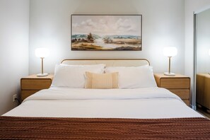 Ein ordentlich bezogenes Bett mit weißen Leinen und einer braunen, strukturierten Decke, an beiden Seiten mit Nachttischen und Lampen, in einem Zimmer mit Landschaftsbild an der Wand.