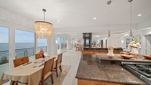 Une cuisine moderne dotée d’un grand îlot central, de luminaires suspendus et offrant une vue sur l’océan.