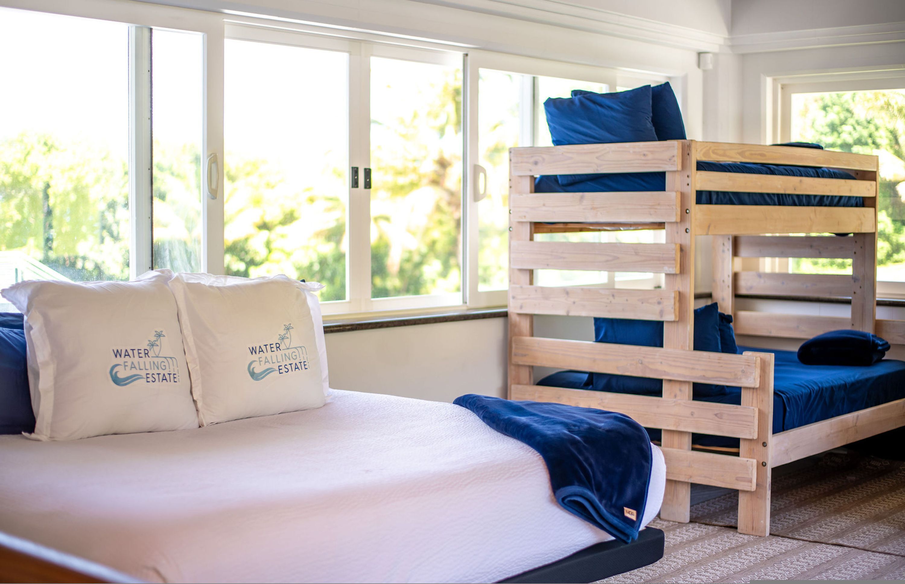 Een stapelbed met houten frame en blauw beddengoed, een enkel bed met wit beddengoed en bijpassende kussens, en grote ramen met uitzicht op groen.