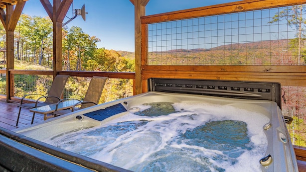 A hot tub with a wooden deck and chairs, surrounded by a wooden fence and a view of a forested landscape.