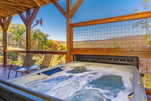 A hot tub with a wooden deck and chairs, surrounded by a wooden fence and a view of a forested landscape.