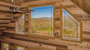 A wooden cabin with a sloped ceiling, a ceiling fan, and large windows offering a view of a mountainous landscape.