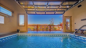An indoor swimming pool with clear blue water, a view of a wooden deck and mountains through a glass ceiling, and a poolside area with a metal ladder and a sign on the wall.