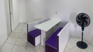 Une petite pièce propre, avec un bureau blanc, des coussins violets, un ventilateur sur pied et un sol carrelé.