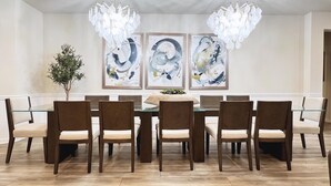 Un comedor con una mesa de vidrio rodeada por seis sillas, una araña y tres pinturas abstractas en la pared.