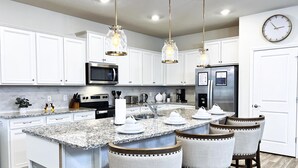 Una cocina moderna con gabinetes blancos, encimera de granito, electrodomésticos de acero inoxidable e iluminación colgante.