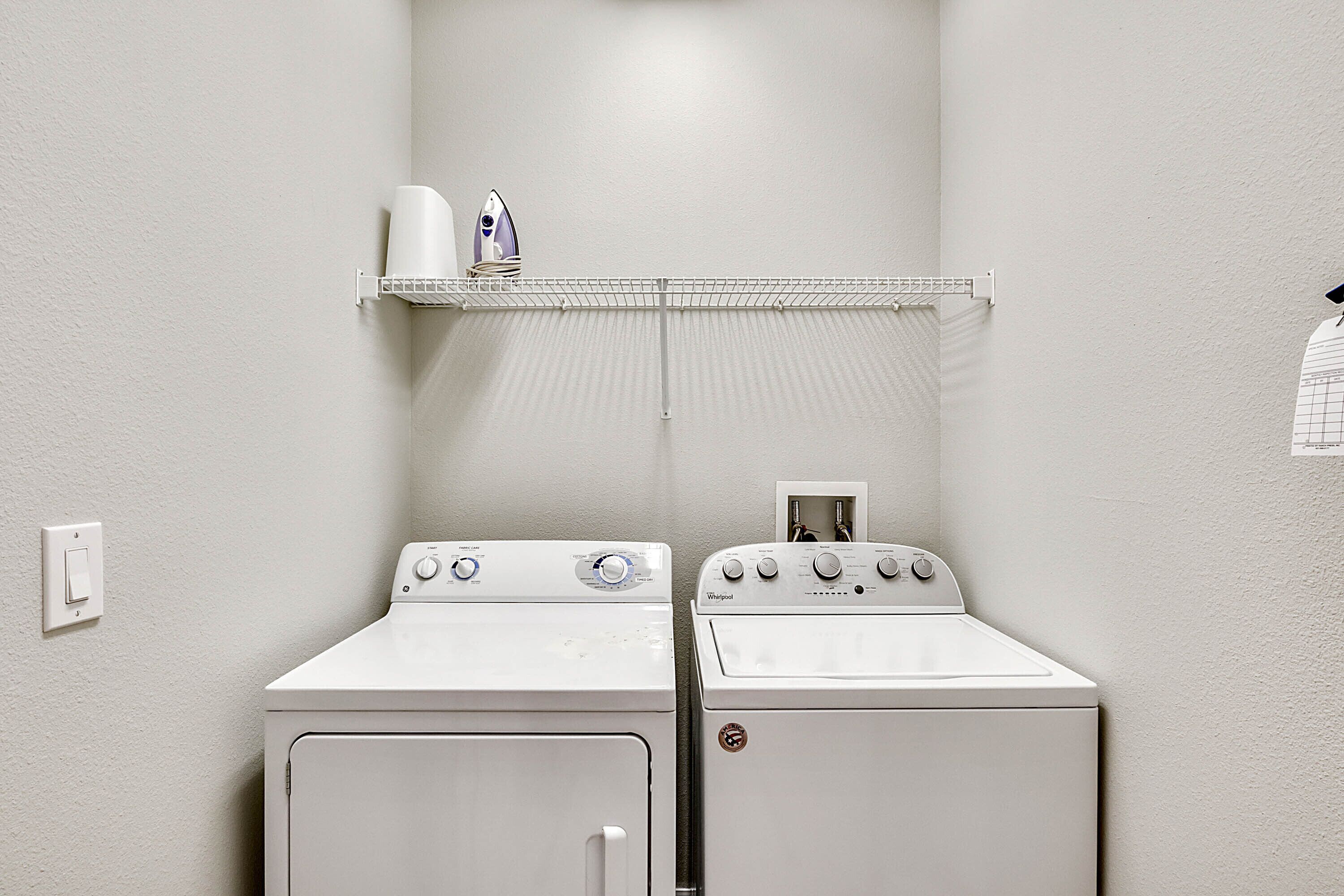 A laundry room with two white washers and dryers, a shelf with a hairdryer and a bottle, and a power outlet on the wall.