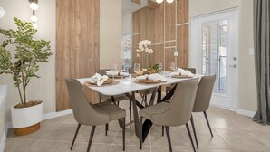 Un coin repas moderne avec une table blanche dressée pour six personnes, entourée de quatre chaises. Un pot de fleurs se trouve à gauche, et un panneau mural en bois, à droite.
