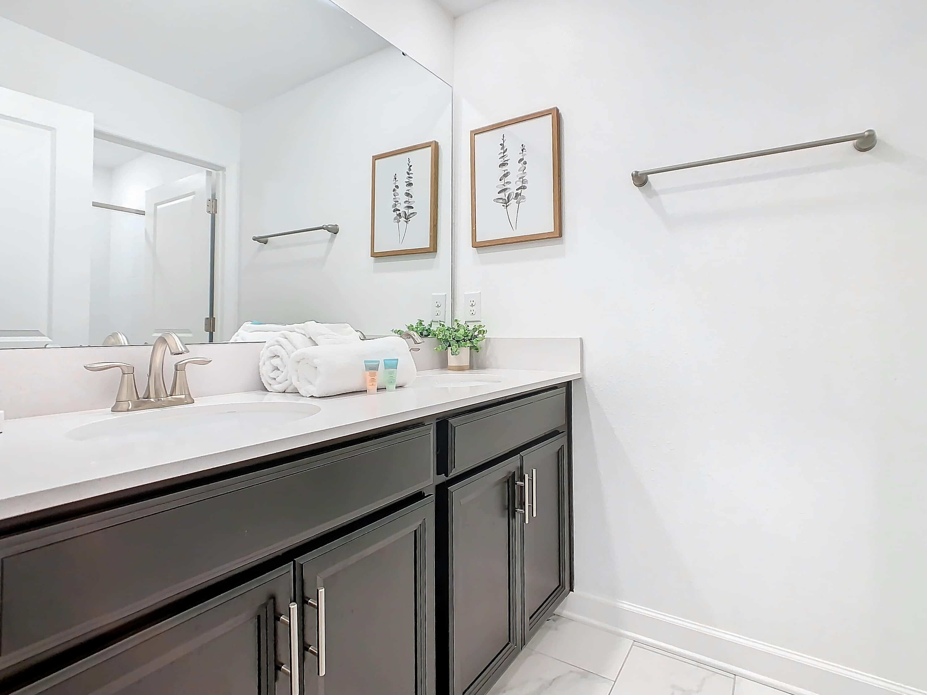 A modern bathroom with a white sink, dark cabinets, and a large mirror. There are towels and toiletries on the counter, and two framed botanical prints on the wall.