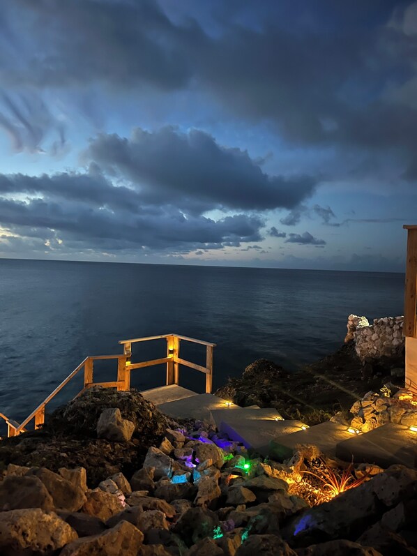 A wooden staircase leading to a rocky platform with colorful lights, overlooking a calm sea under a cloudy sky.