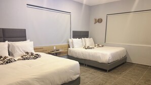 Hotellihuoneessa on kaksi sänkyä, joissa on valkoiset lakanat ja koristeelliset tyynyt. Toisessa sängyssä on puinen pääty ja toisessa harmaa. Sänkyjen välissä on pieni puinen yöpöytä, jossa on kello.