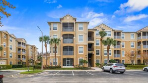 A multi-story residential building with balconies, a parking lot, and palm trees.