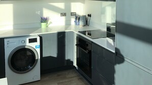 A modern kitchen with dark cabinets, a built-in washing machine, and a countertop with a coffee maker and kettle. - Central Trafford Apartment Free Parking (Manchester)