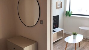 A modern living room with a minimalist design, featuring a white cabinet, a round mirror, a television, and a small table with a plant. - Central Trafford Apartment Free Parking (Manchester)