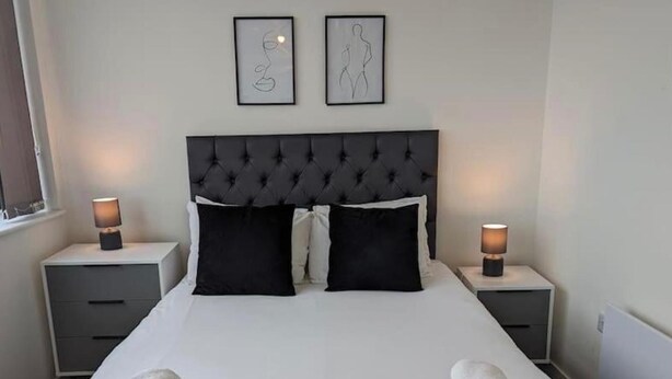 A neatly made bed with two pillows, two bedside tables with lamps, and two framed artworks on the wall. - Central Trafford Apartment Free Parking (Manchester)