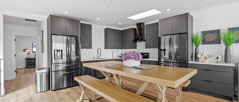 A modern kitchen with grey cabinetry, stainless steel appliances, a wooden dining table, and a bench.