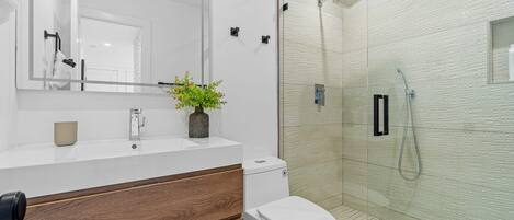 A modern bathroom with a white vanity, wooden drawer, white toilet, and a glass shower enclosure.