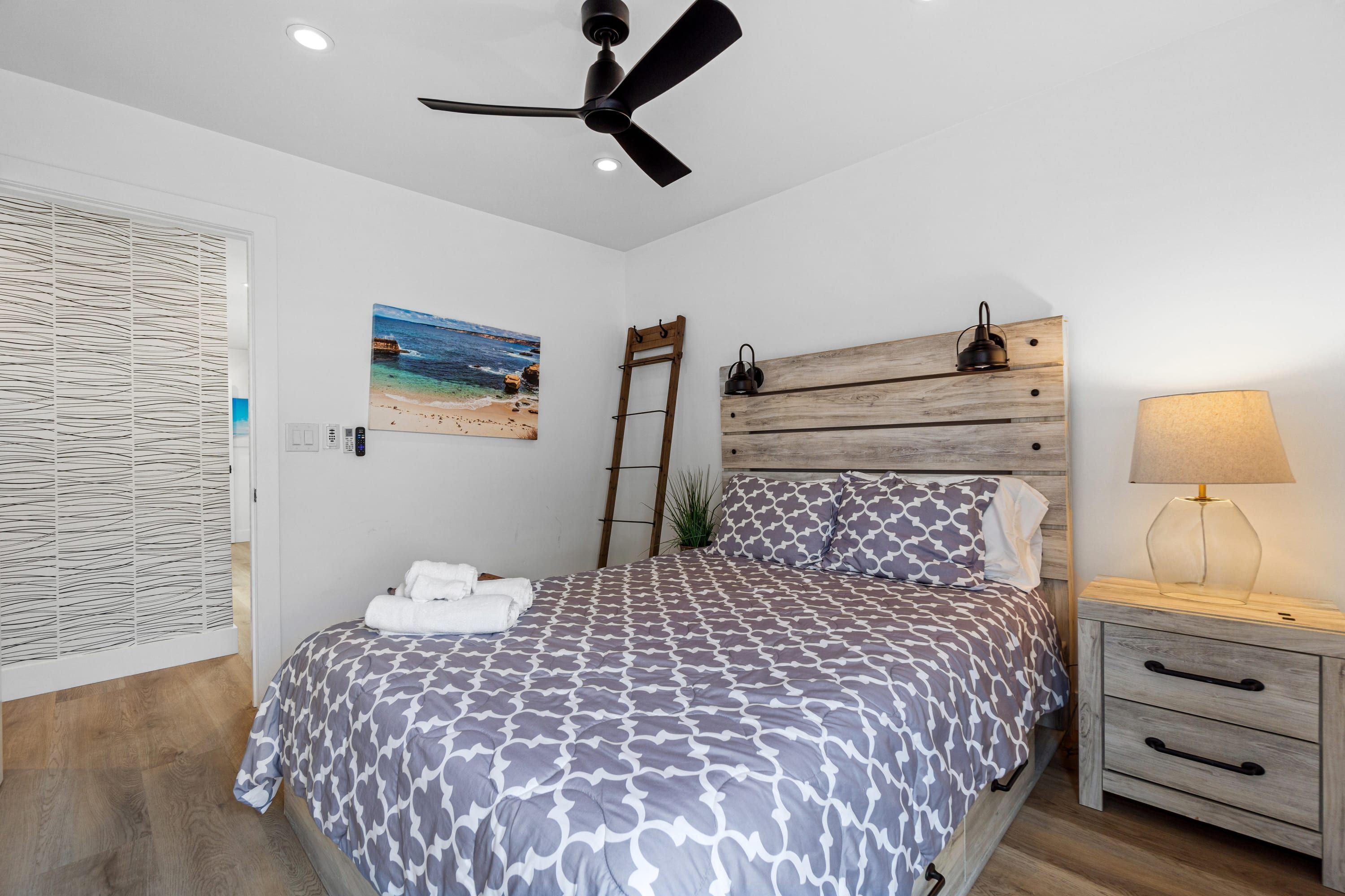 A bedroom with a bed, a ceiling fan, a wooden headboard, a bedside table with a lamp, and a painting on the wall.